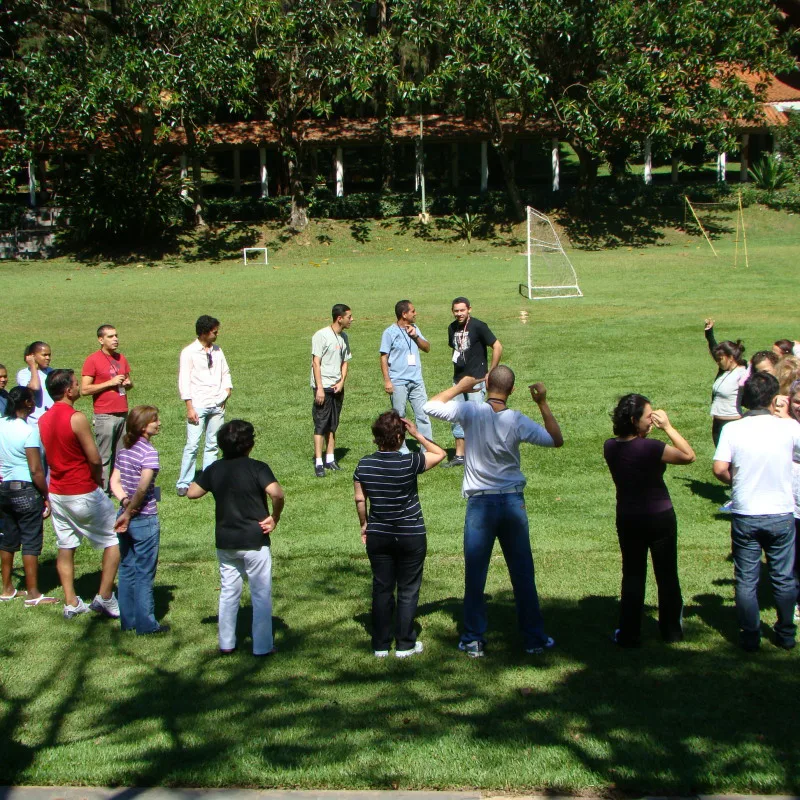 Dinâmica de grupo ao ar livre: quinze pessoas em círculo num gramado participando de atividade cooperativa